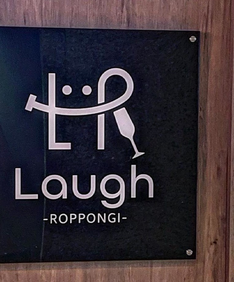 木目の壁に取り付けられた黒いアクリルの看板。白い文字で「Laugh -ROPPONGI-」とロゴマーク。